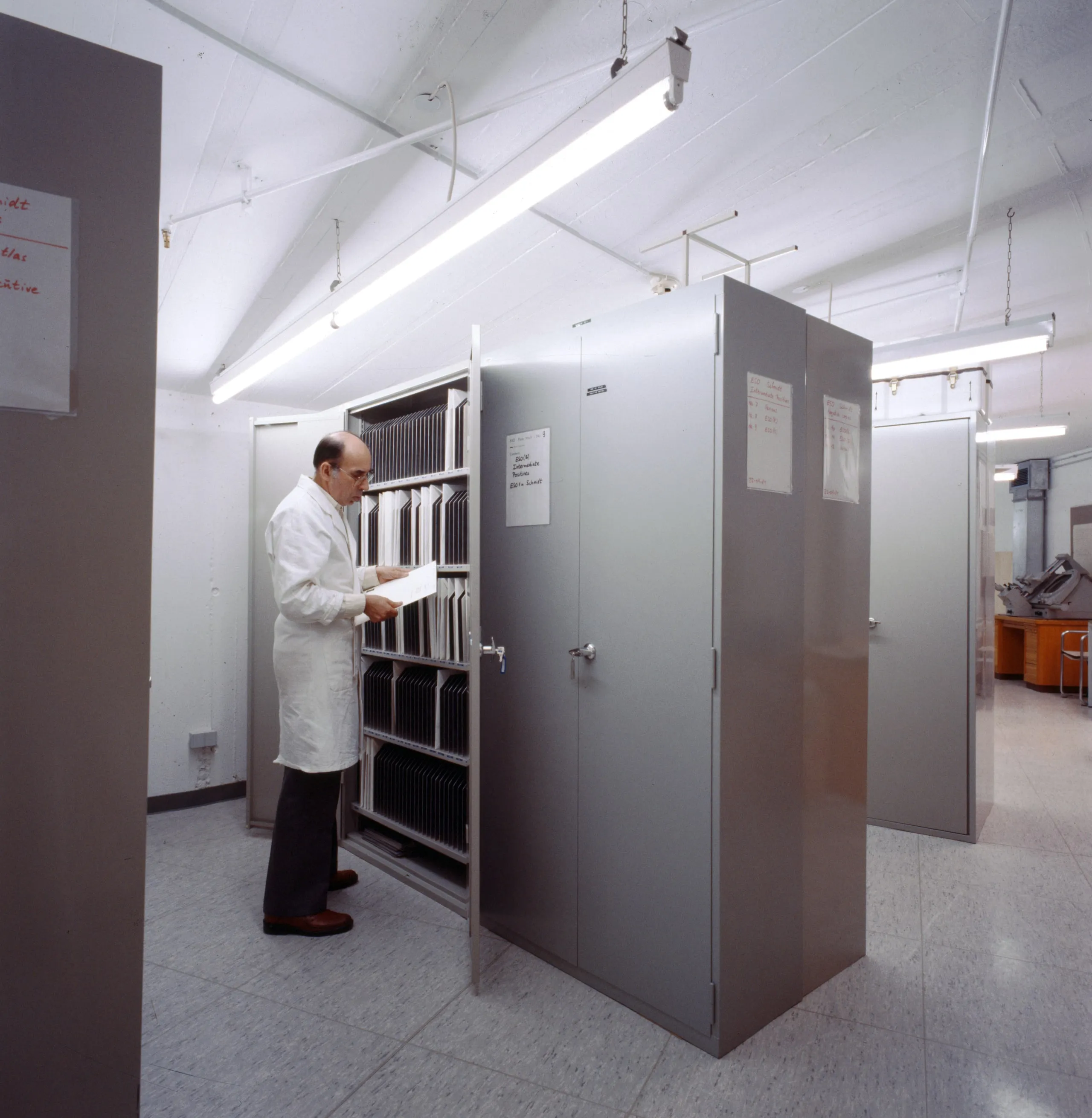 A man in a white lab coat reading a sheet beside open grey metal cabinets filled with rows of photographic plates.