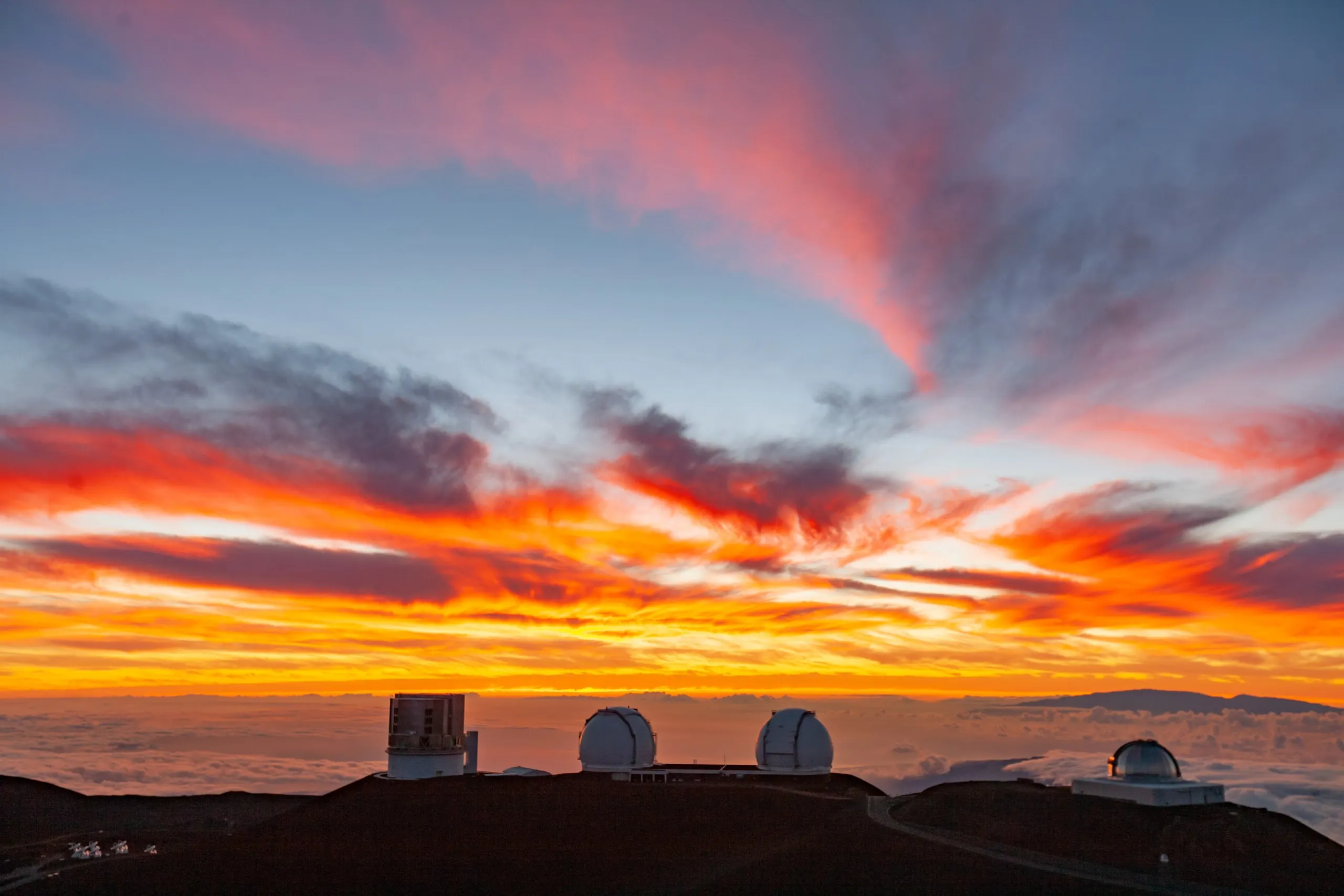 Observatory domes on the summit ridge of Maunakea under an orange and red sunset sky, above a layer of clouds.