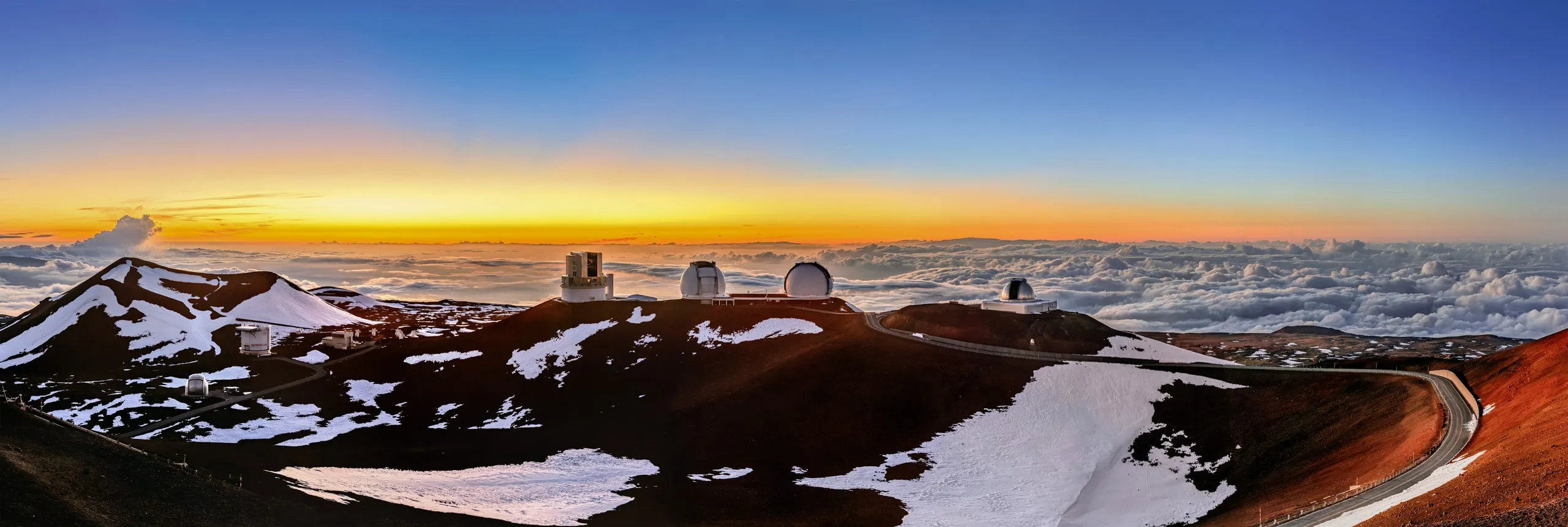 Snow-dusted red cinder slopes and telescope domes on Maunakea at sunset, with clouds below.