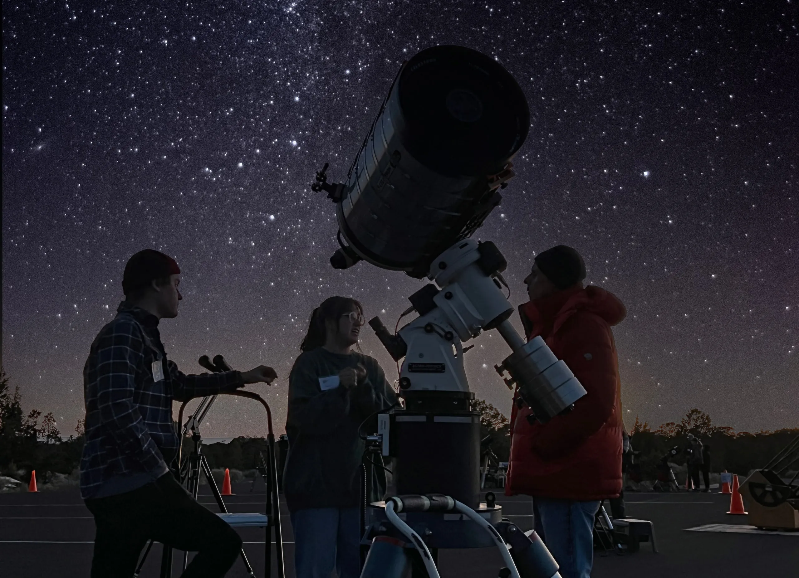 Three people in silhouette around a large amateur telescope on a mount under a starry sky.